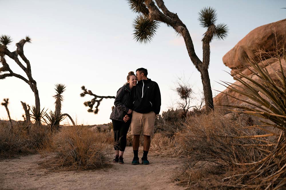 A smiling couple stands together on a desert path at dusk, surrounded by Joshua trees and large rock formations.