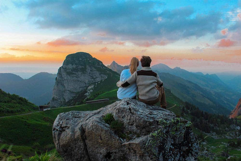 A couple sitting on a rock facing a mountain view in a natural outdoor setting.